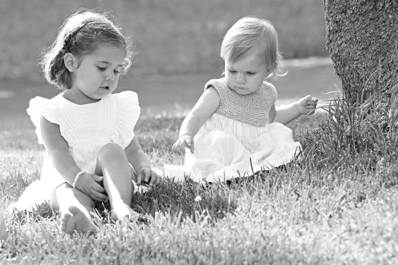 Dos niñas pequeñas, vestidas de blanco, juegan en la hierba junto a un árbol.